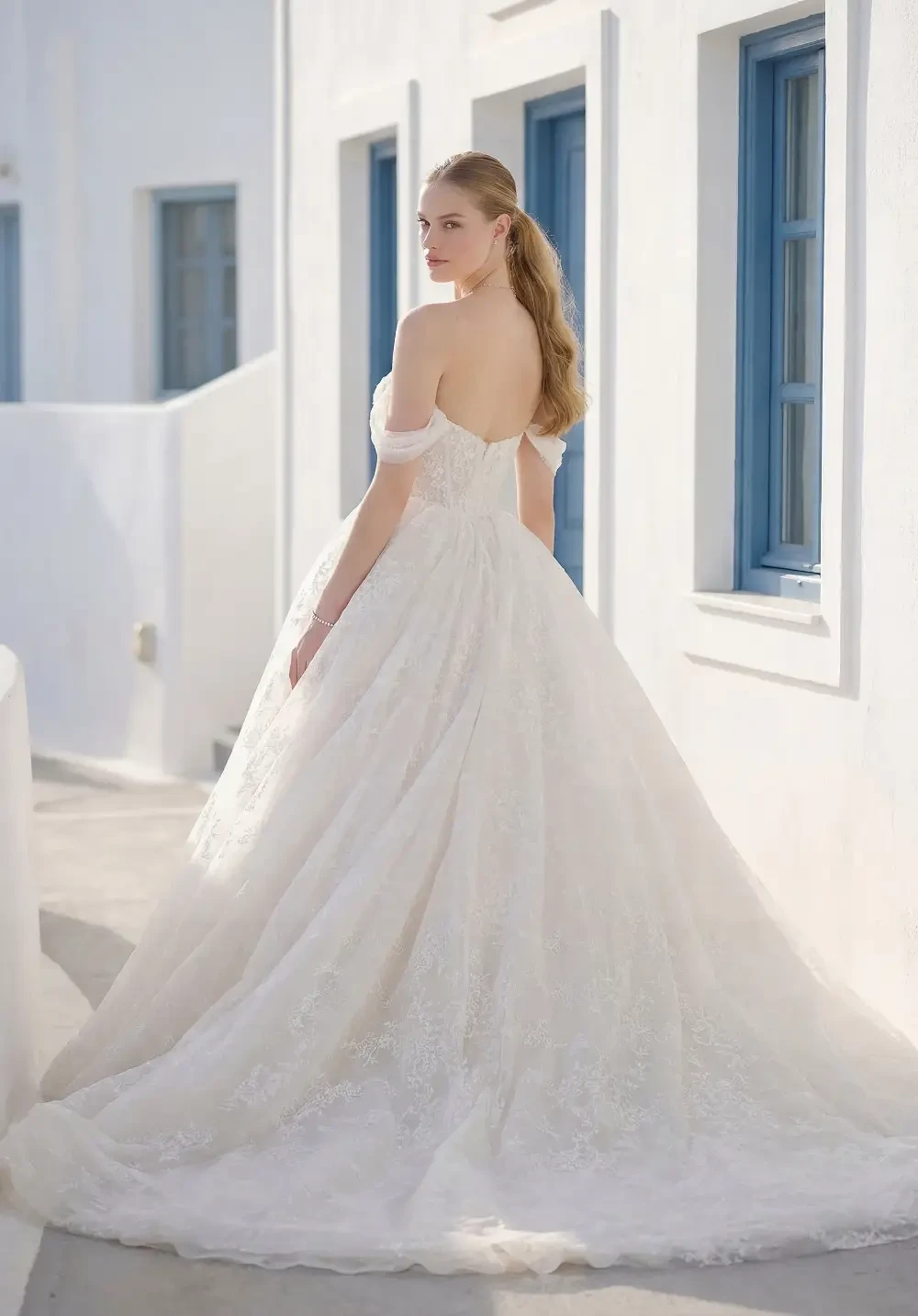 A woman in a wedding dress stands with her back to the camera, looking over her shoulder. The dress is white and has a flowing train. She is in a light, airy setting with white walls and blue windows.