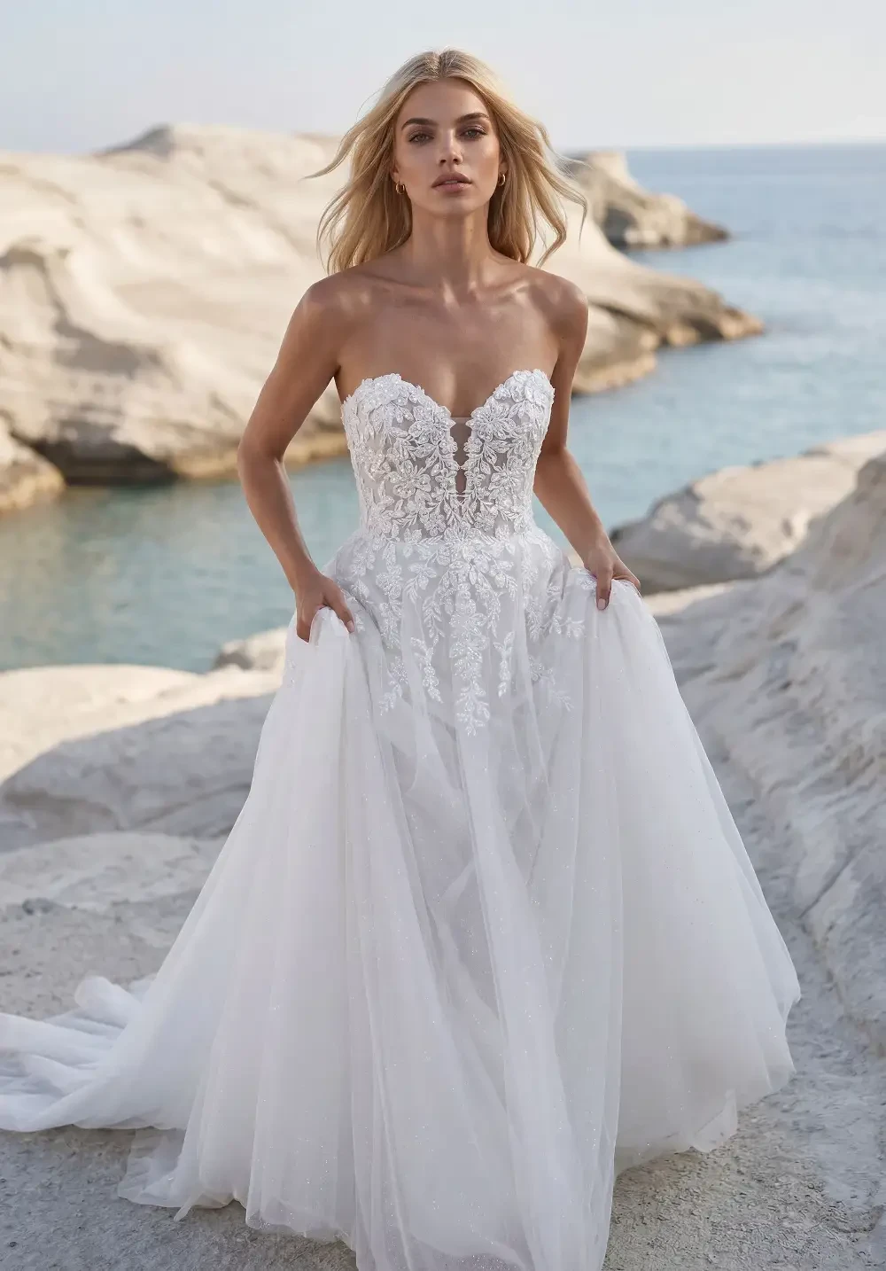 A woman wearing a strapless, lace wedding dress stands on a rocky shore near the ocean.