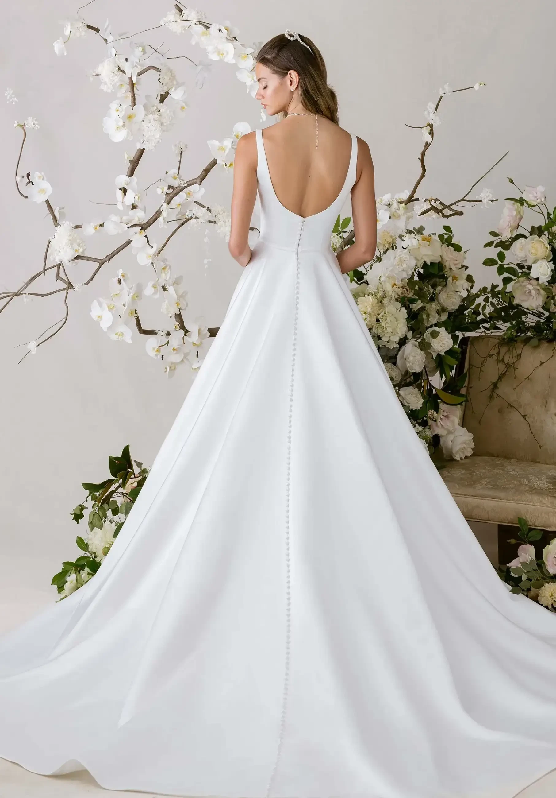 A woman wearing a simple, elegant white wedding dress with a low back stands in front of floral arrangements and branches.