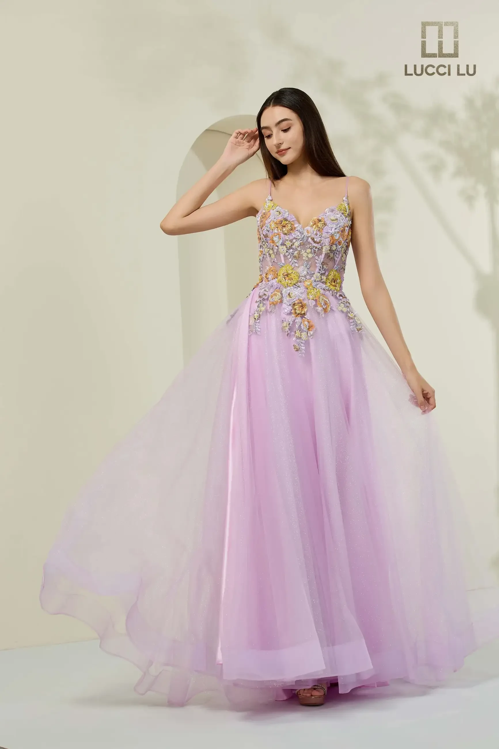 A woman in a flowing lavender gown adorned with intricate floral embellishments poses gracefully in a soft, light-filled space, exuding elegance.