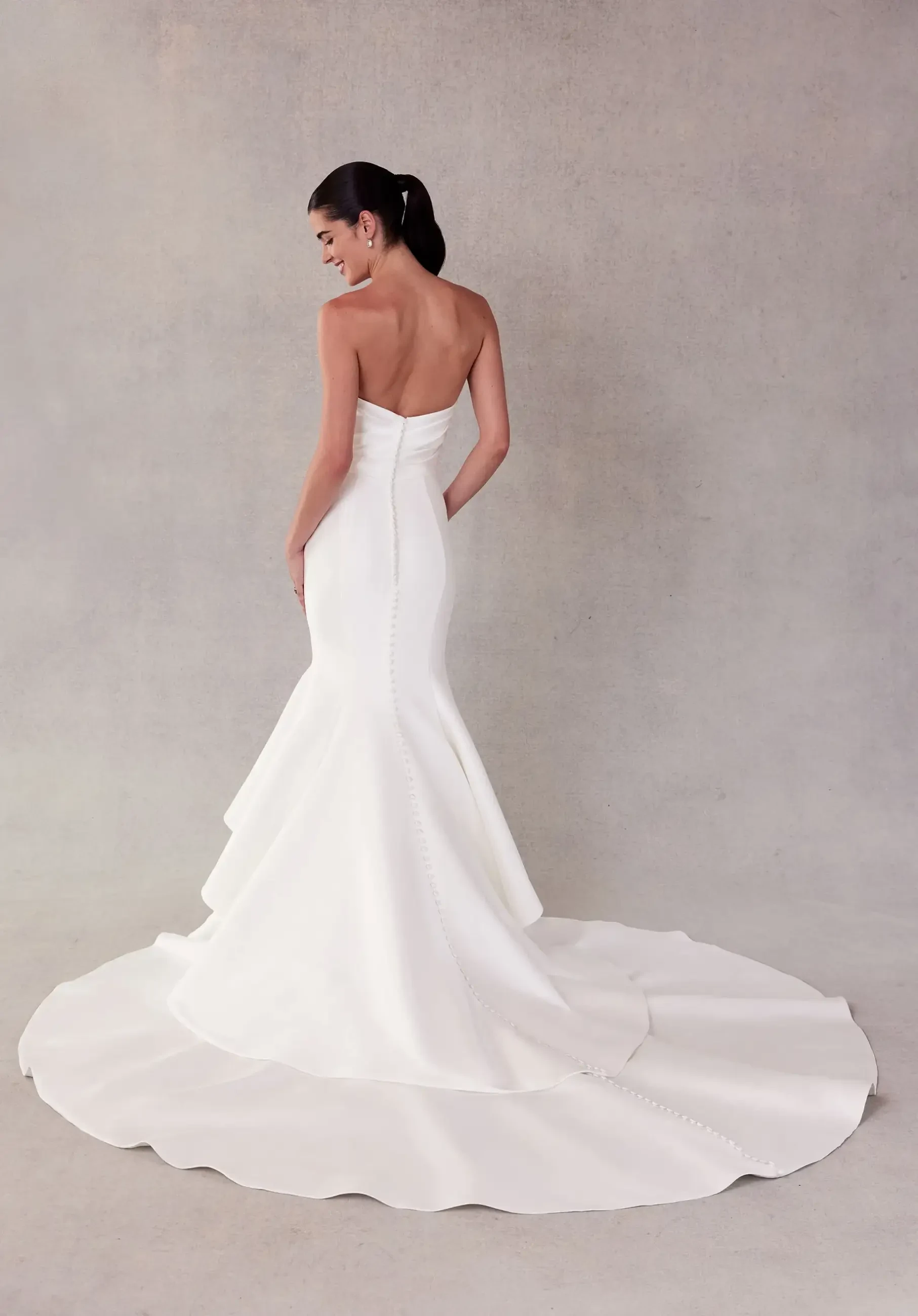 Model is wearing a strapless satin mermaid gown with a draped cowl bodice, tiered skirt, and buttons running to the hemline.
