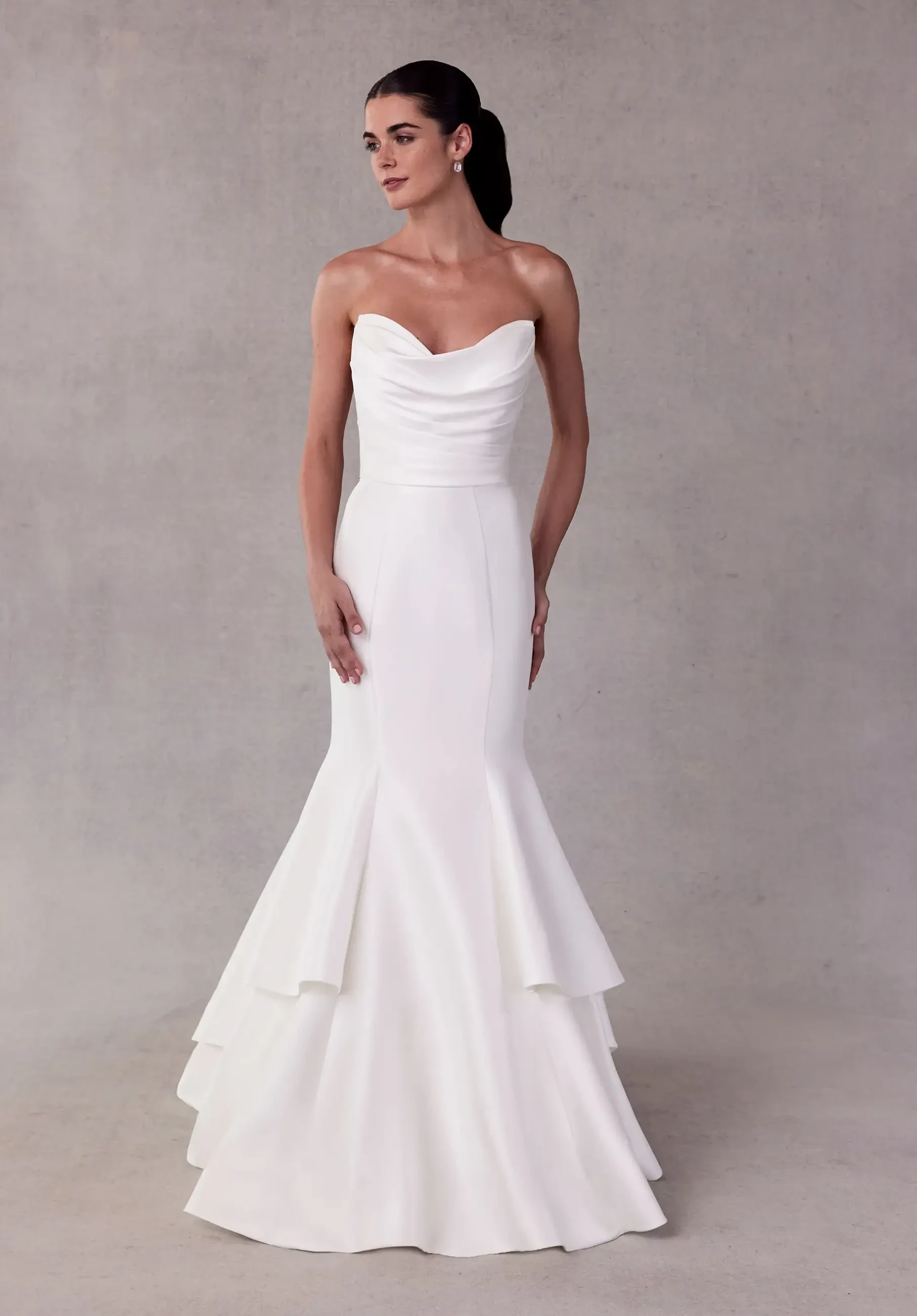 Model is wearing a strapless satin mermaid gown with a draped cowl bodice, tiered skirt, and buttons running to the hemline.