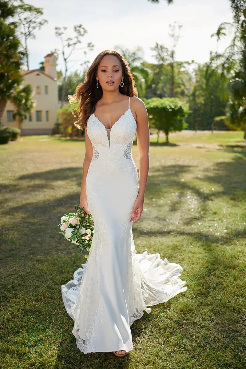 Bride in a fitted, lace wedding dress holding a bouquet, standing on grassy field. Sunlight creates a serene mood, with trees and a house in the background.