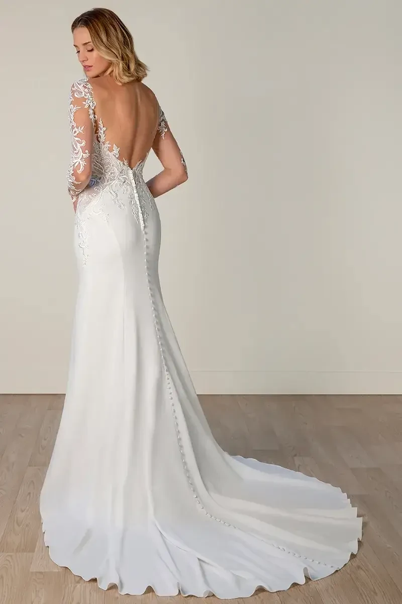 Model is wearing a lace fit-and-flare gown with a high illusion neckline, sheer long lace sleeves, a low open back, and a sleek trumpet skirt.