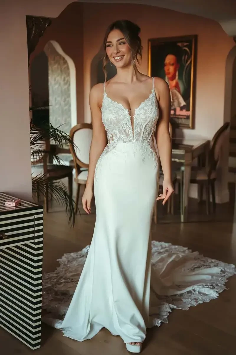 A woman is standing and smiling in a fitted wedding dress with intricate lace details. The dress has thin straps and a deep neckline, complemented by a flowing train. She is in an elegantly decorated room with soft lighting, plants, and furniture visible in the background.